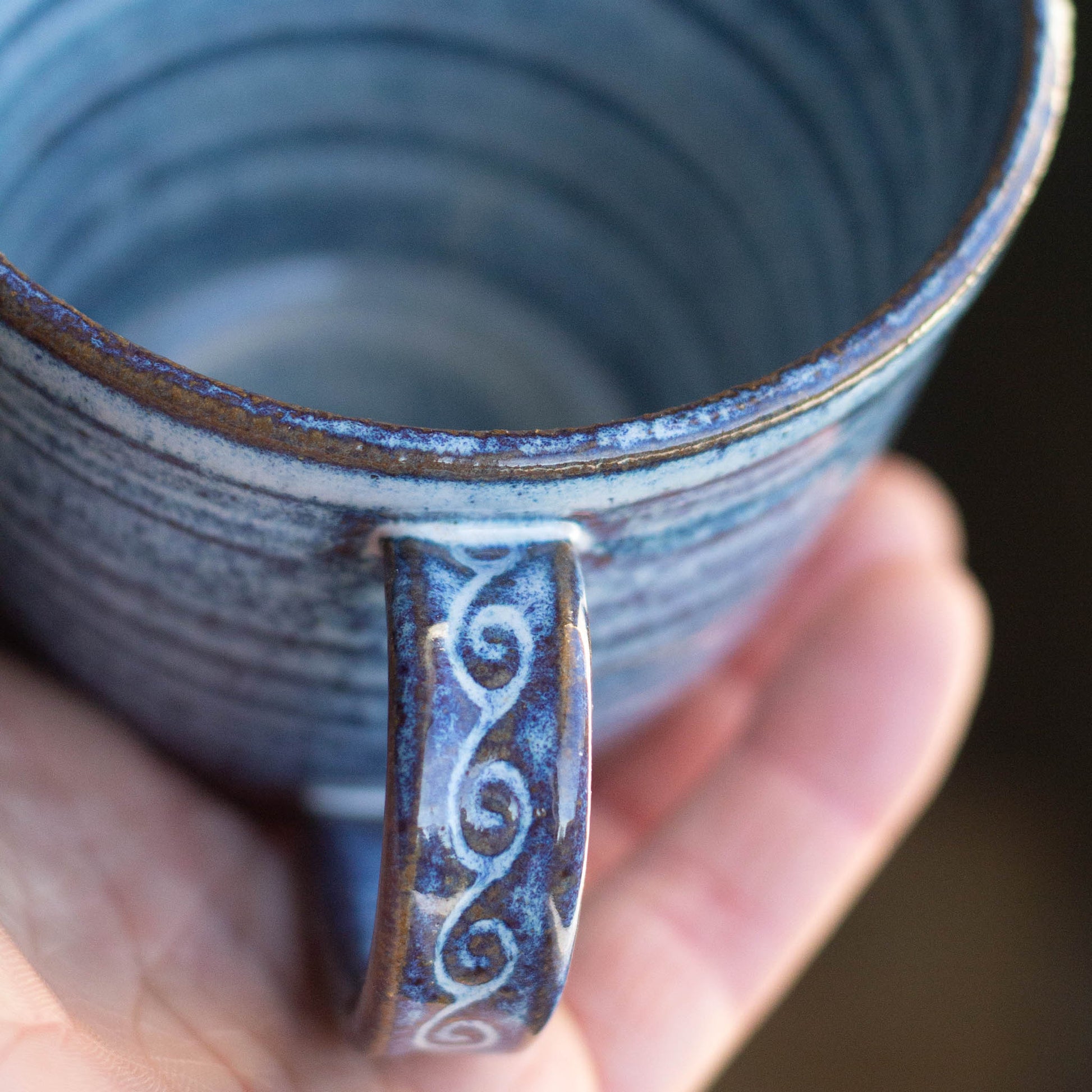 Hand holding a blue ceramic mug with swirling hnadthrown patterns and decorative handle