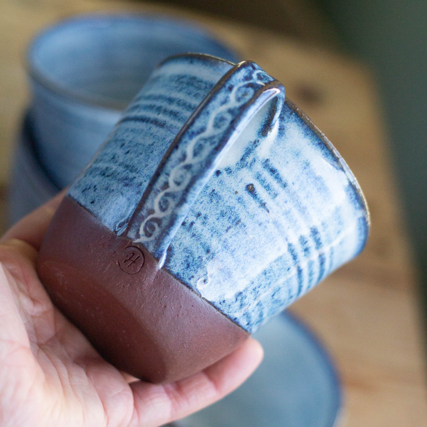Hand holding a blue ceramic cup with red brown base and decorative handle on a wooden surface