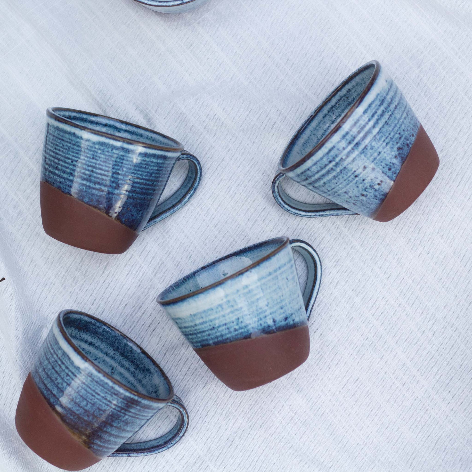 Four ceramic mugs with blue glaze and red brown bases on a white textured surface