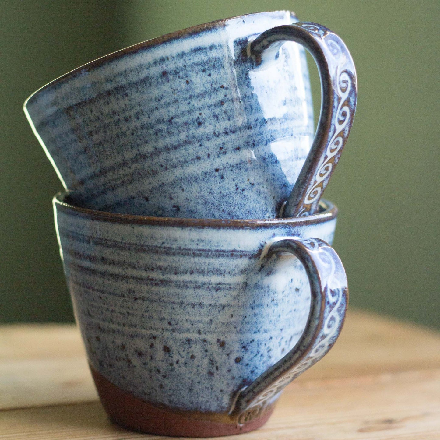Two blue ceramic mugs with decorative handles stacked on a wooden surface.