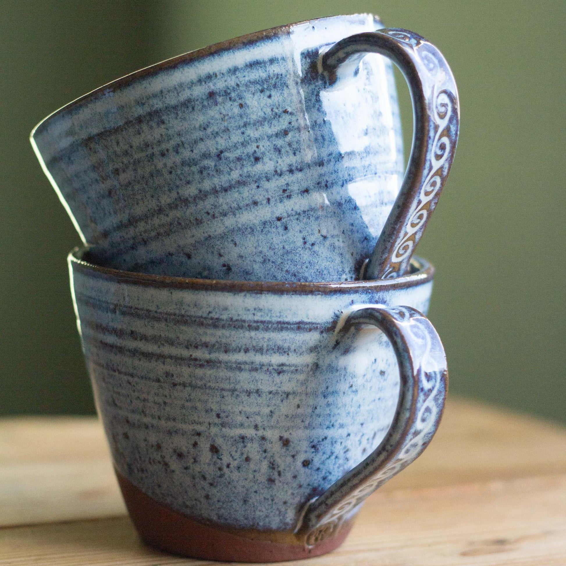 Two blue ceramic mugs with decorative handles stacked on a wooden surface.